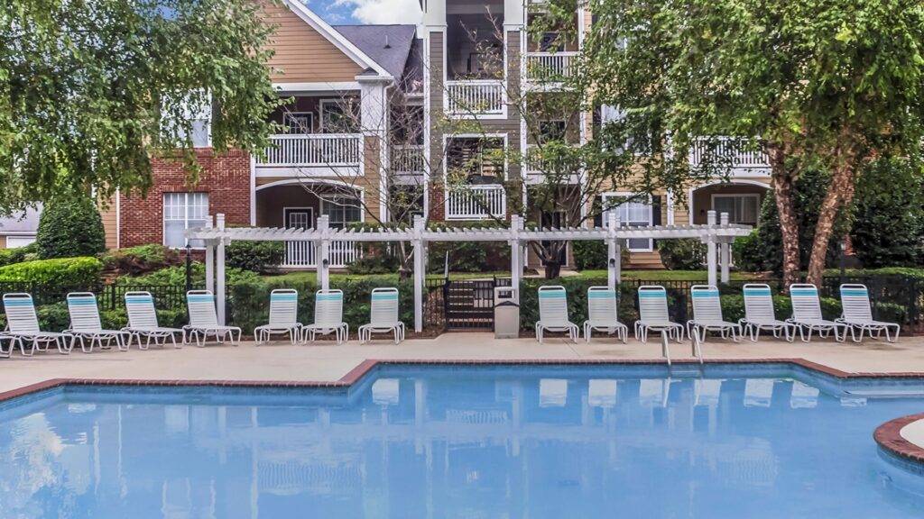 A wide-angle view of a sparkling blue resort-style swimming pool at The Legends, featuring a long row of white lounge chairs under a white pergola with modern apartment buildings