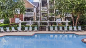 A wide-angle view of a sparkling blue resort-style swimming pool at The Legends, featuring a long row of white lounge chairs under a white pergola with modern apartment buildings